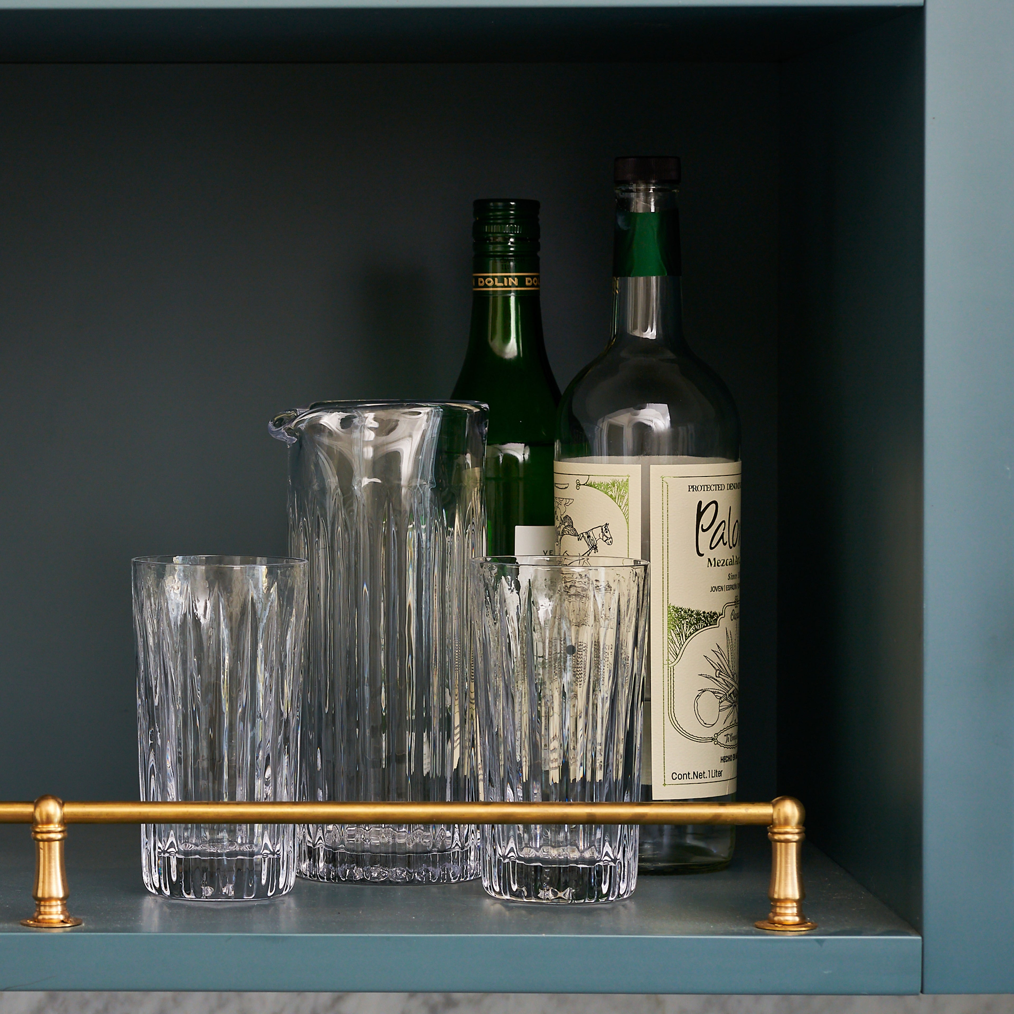 Two clear glass highball glasses with vertical ridge pattern, matching pitcher, and two bottles displayed on shelf.