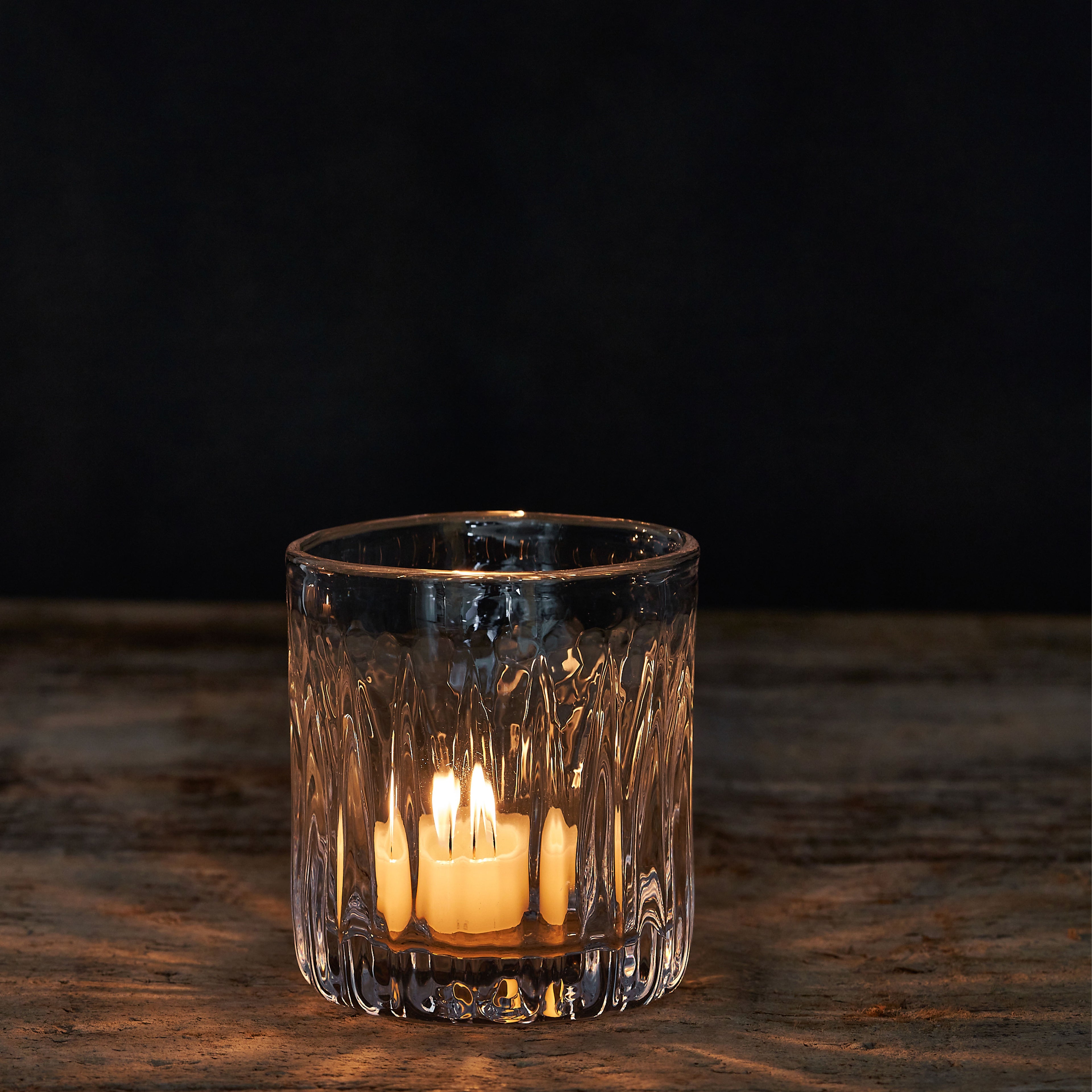 Clear glass tealight holder with textured ridged pattern and lit cream-colored tealight candle inside.