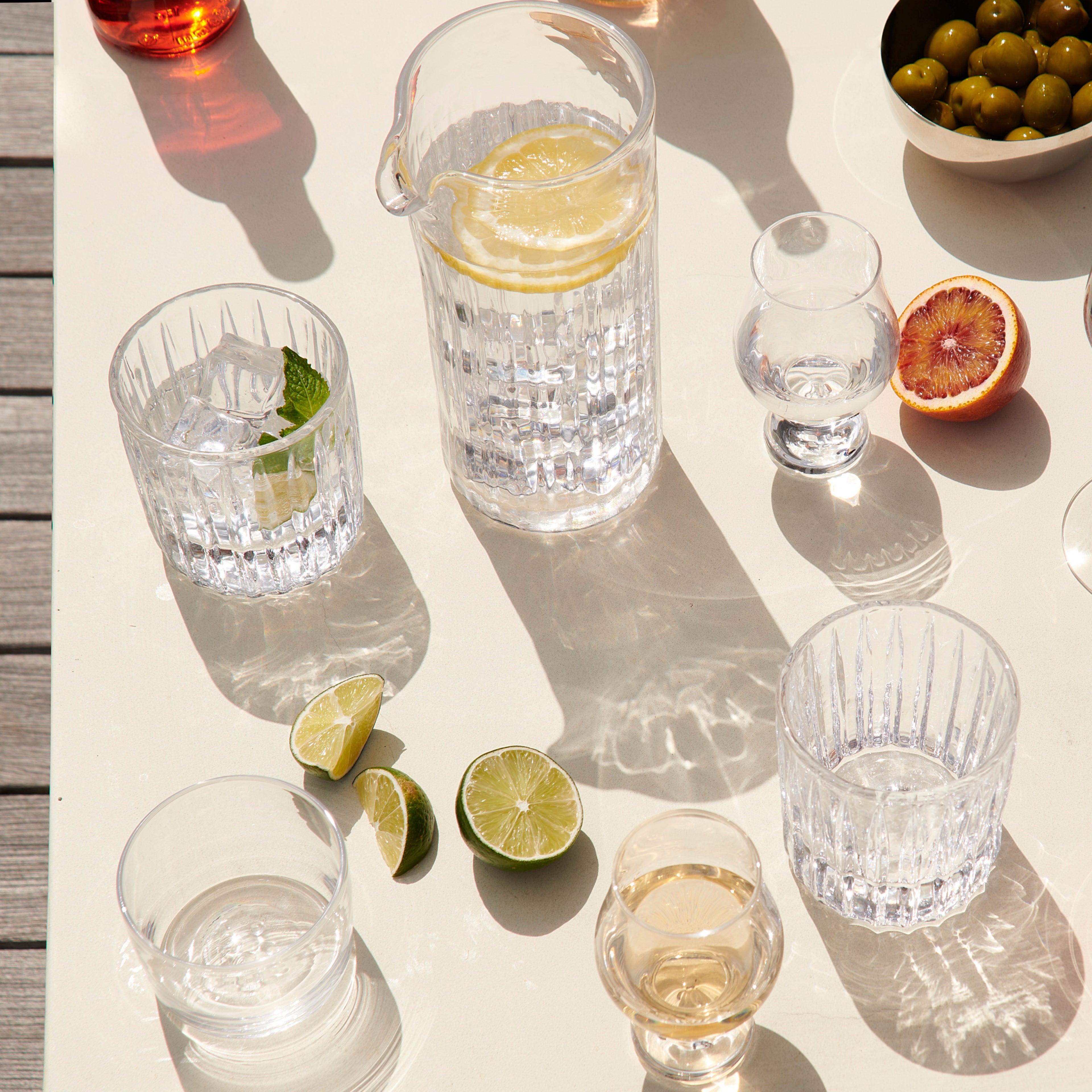 Tall clear glass pitcher with lemon slices and water, surrounded by matching textured glasses and citrus fruit.