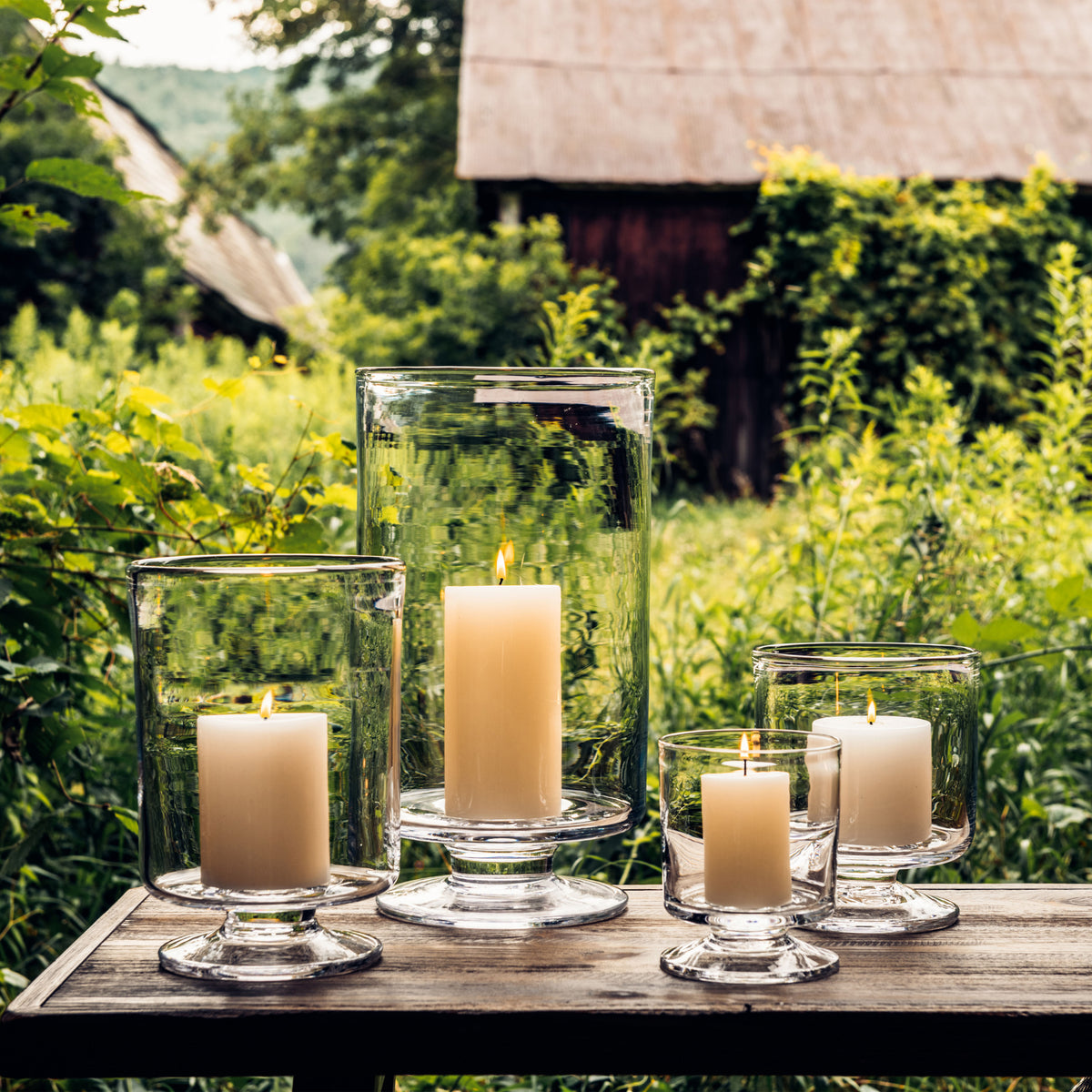Four glass hurricane candle holders of varying heights with cream pillar candles inside, arranged on wooden surface.
