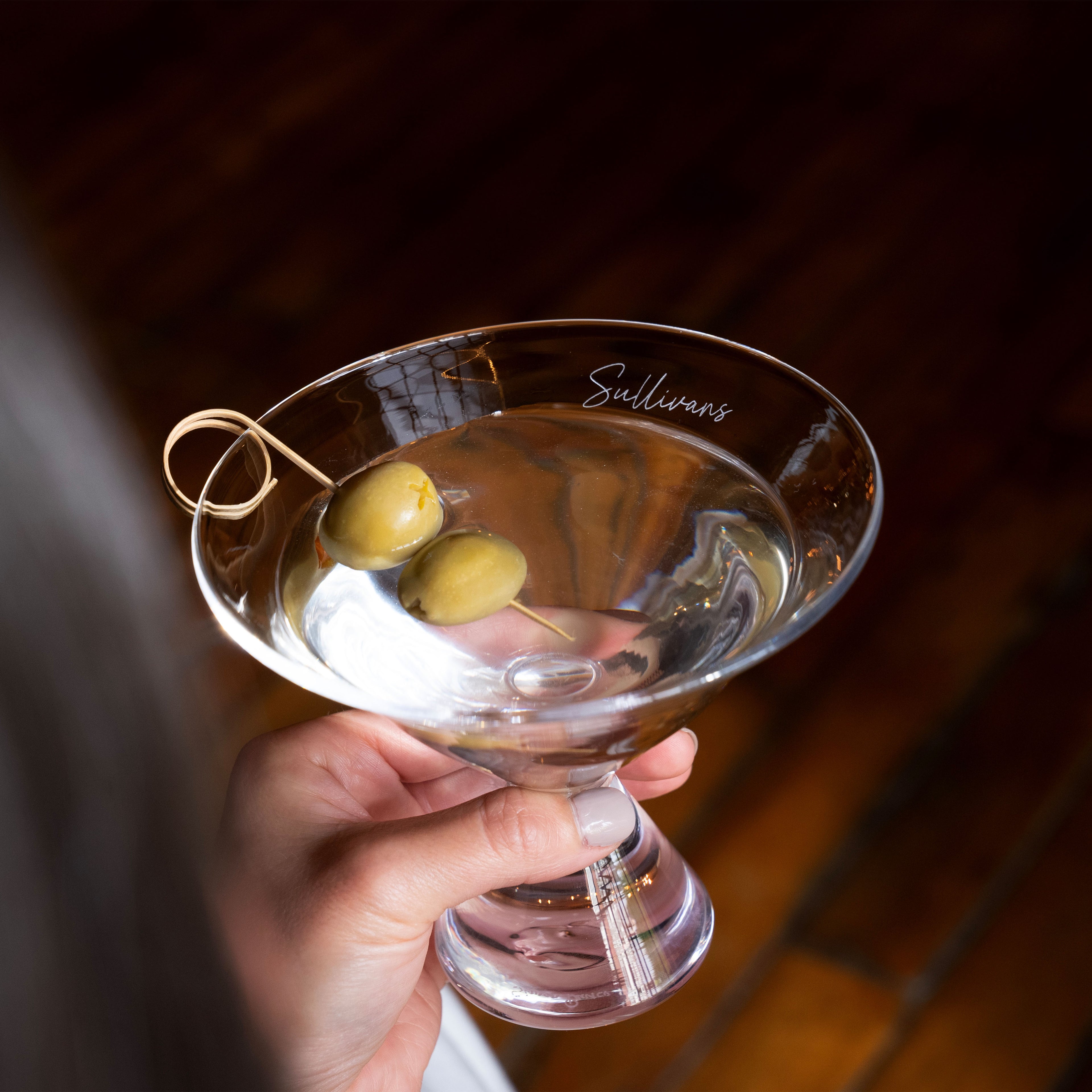 Hand holding a martini glass filled with clear liquid and two green olives on a pick, with "Sullivans" engraved on the rim.