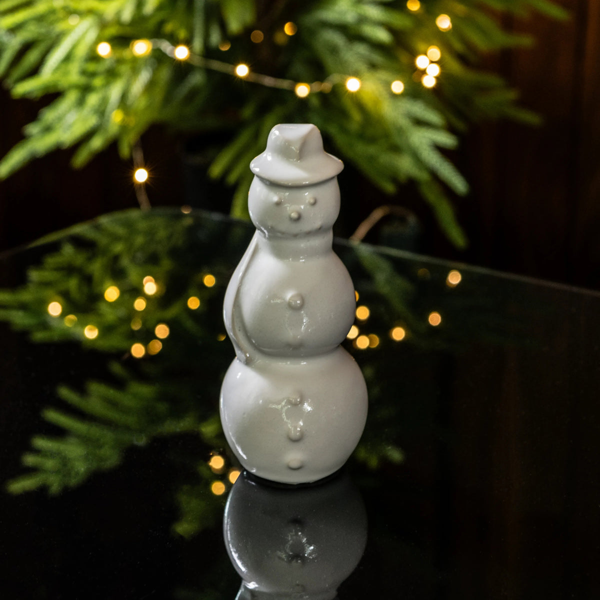 White ceramic snowman figurine with stacked body, coal buttons, and hat, displayed among evergreen branches and warm lights.