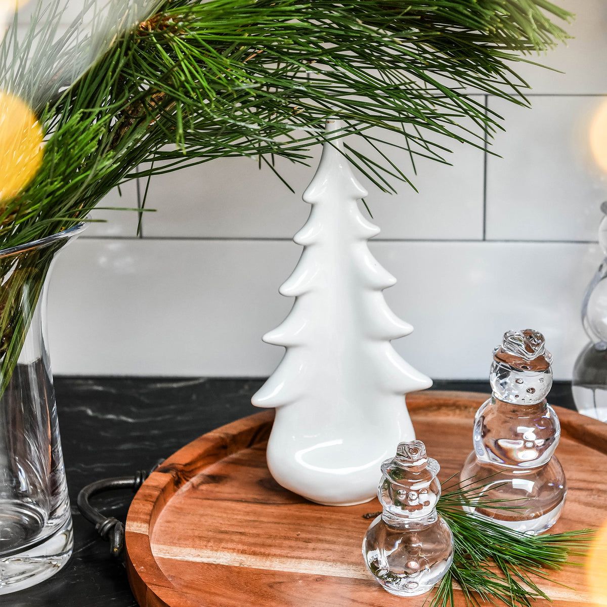White ceramic tree-shaped ornament displayed on wooden tray with fresh evergreen branches and glass accents.