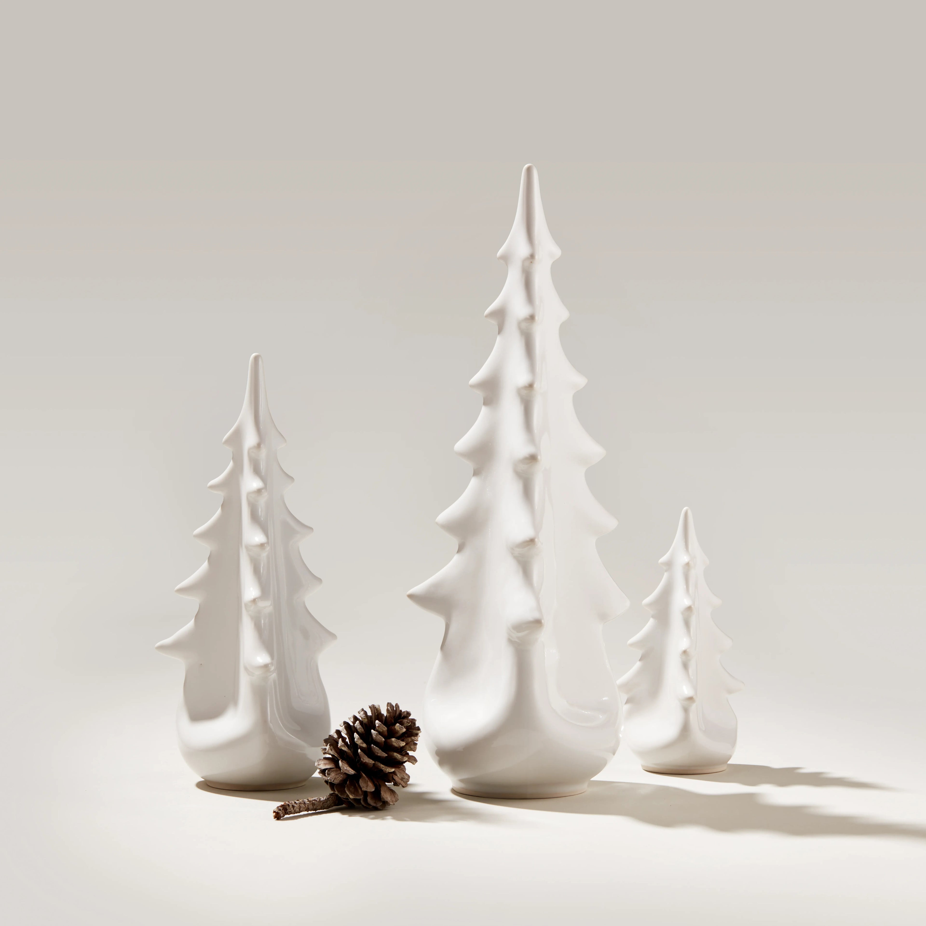 White ceramic evergreen tree figurines in three graduated sizes with a pinecone.