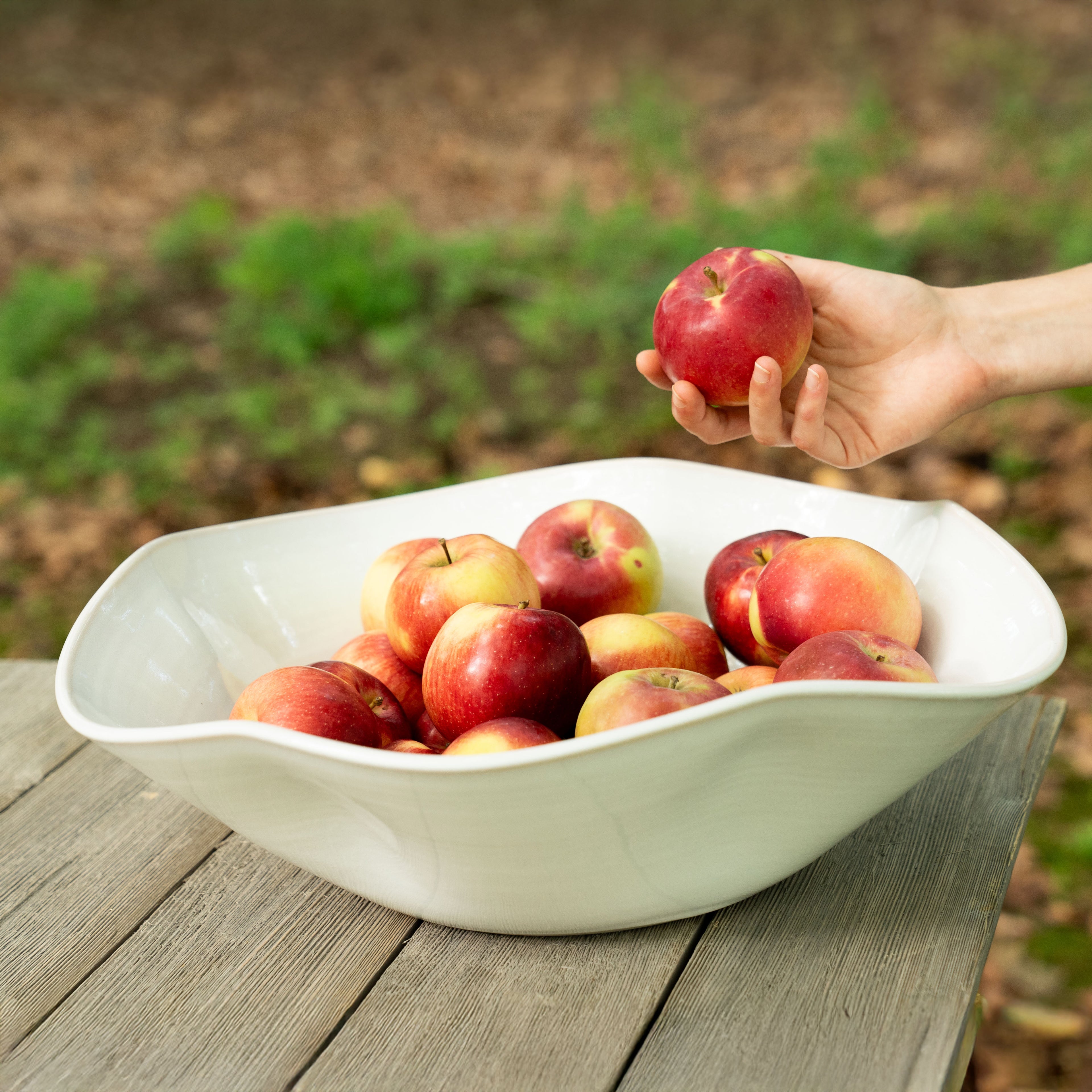 White curved centerpiece bowl filled with red apples, with one apple held above by a hand.