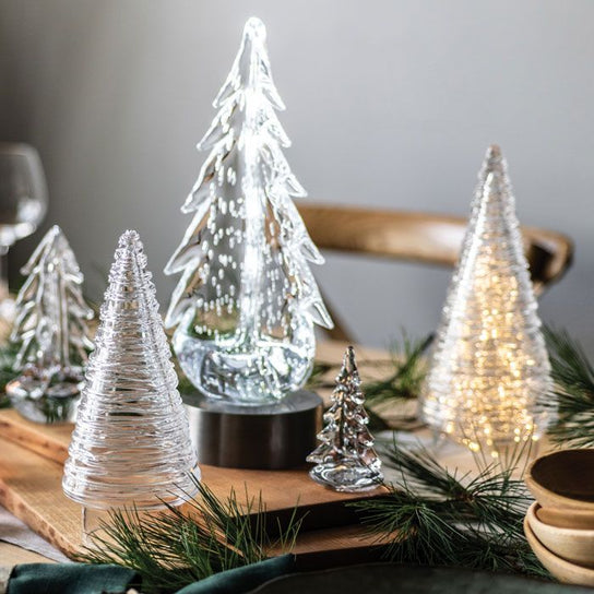 Decorative illuminated glass and metallic tree figurines in various sizes arranged on a wooden tray with greenery.