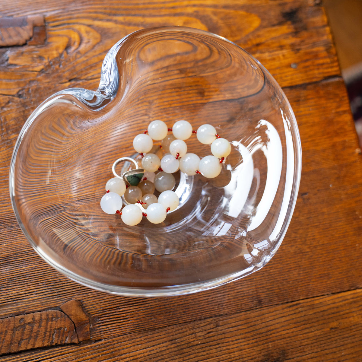 Clear glass heart-shaped dish containing white and tan beaded bracelet with gold accents, sitting on wood surface.