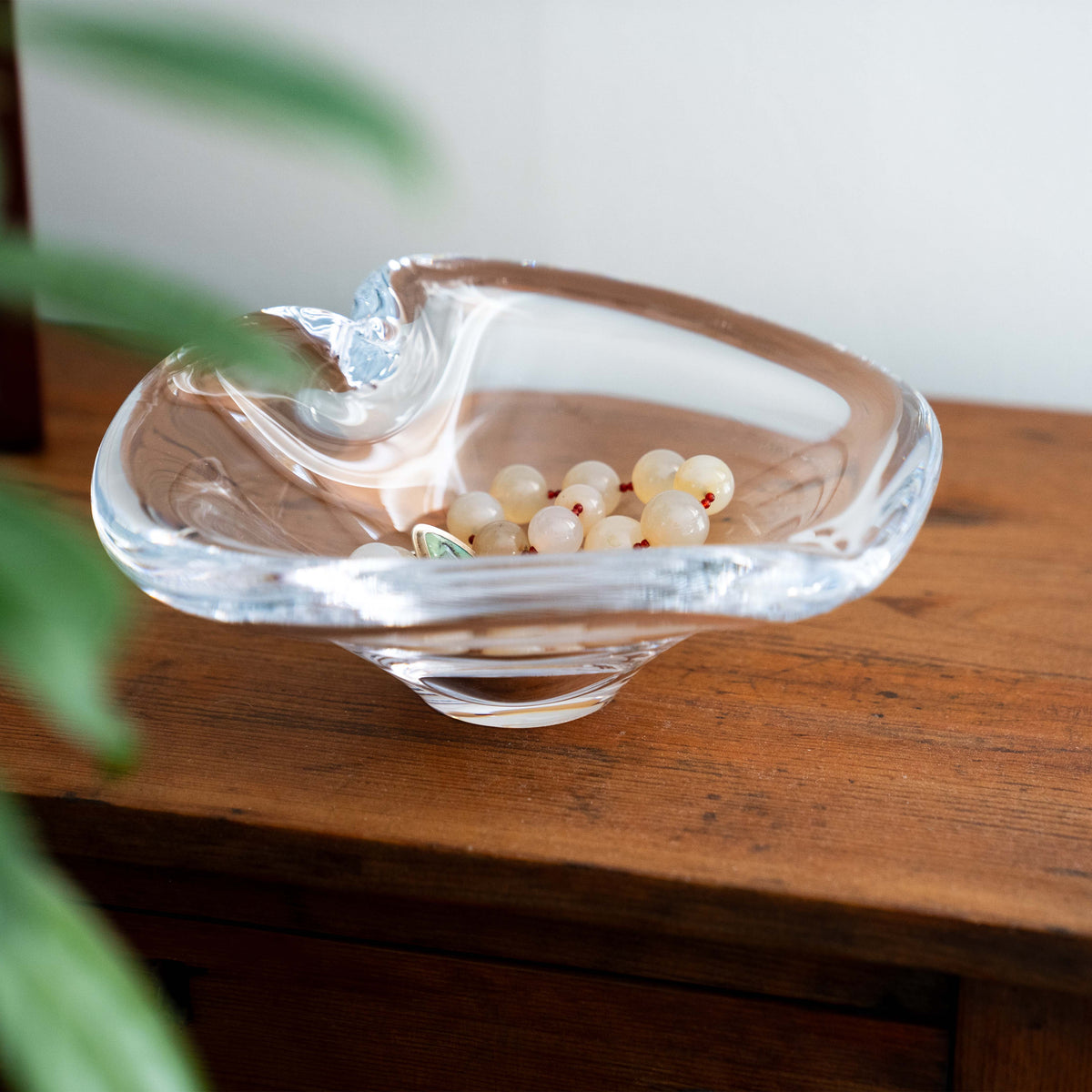 Clear glass heart-shaped dish holding small cream and red beads, displayed on a wooden surface.