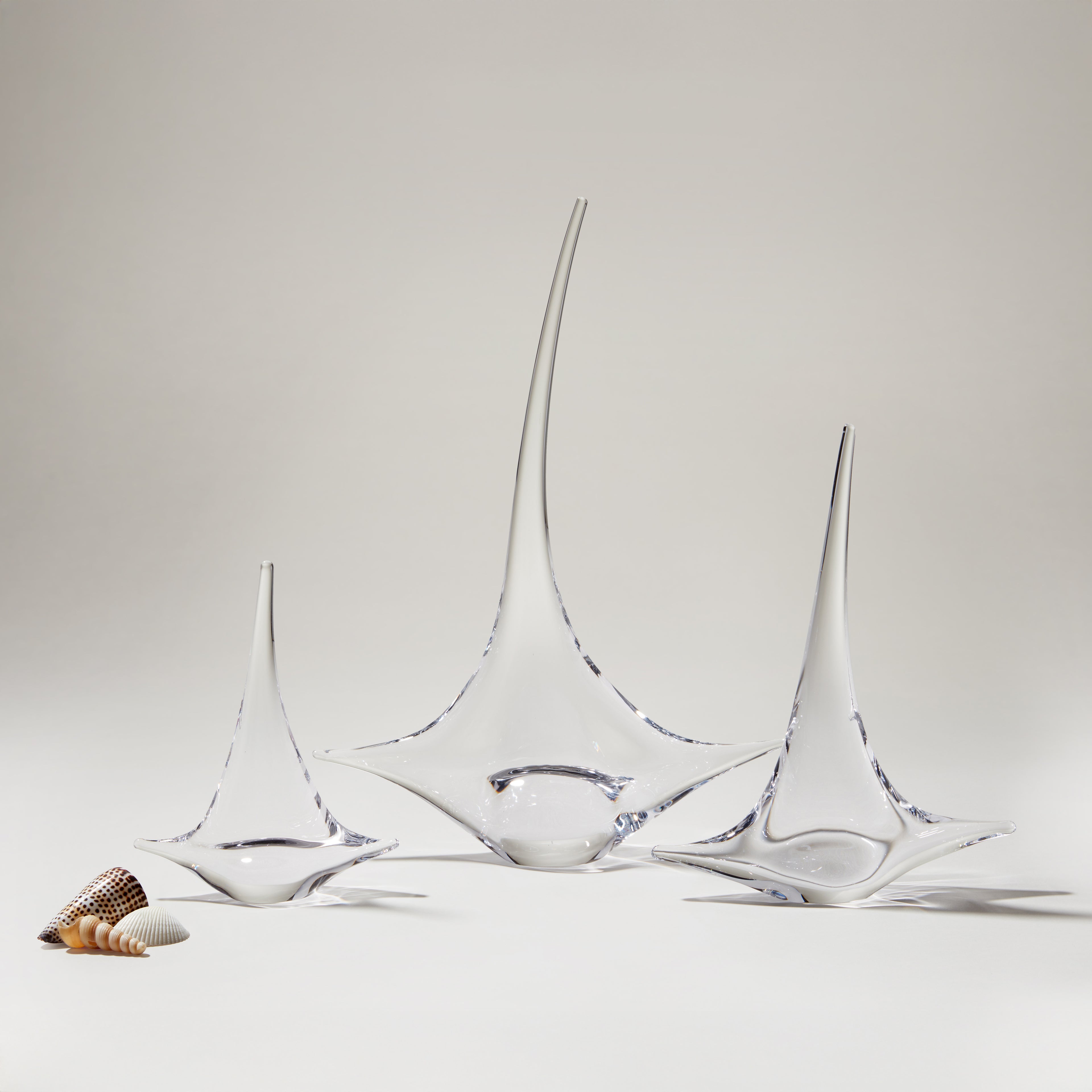Three glass sailboat figurines with blue accents, displayed with small shells on white surface.