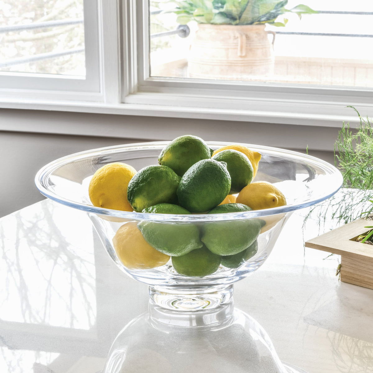 Clear glass pedestal bowl filled with green and yellow limes on a white windowsill.