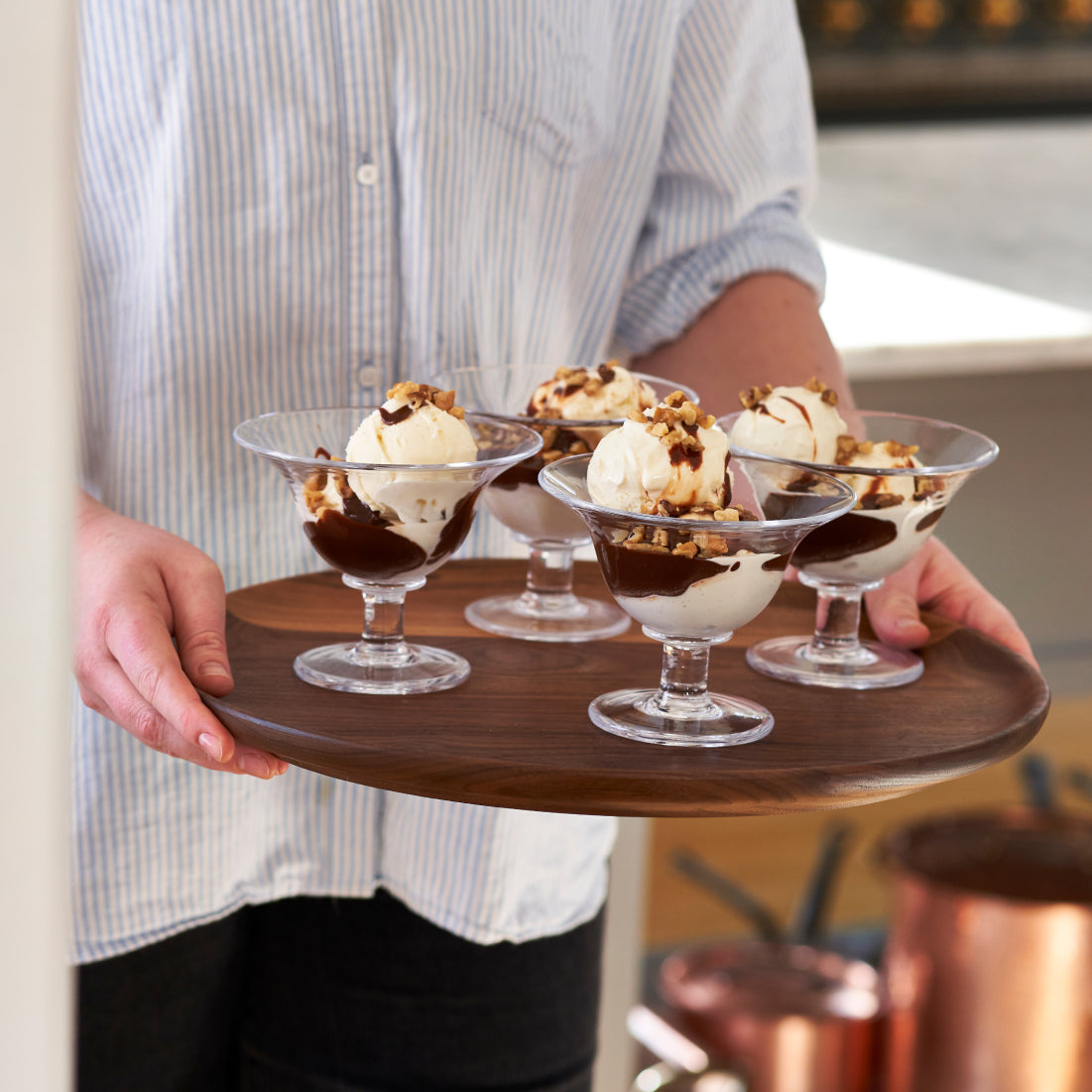 Two clear glass dessert bowls with pedestal bases filled with ice cream and toppings, held on a wooden serving board.