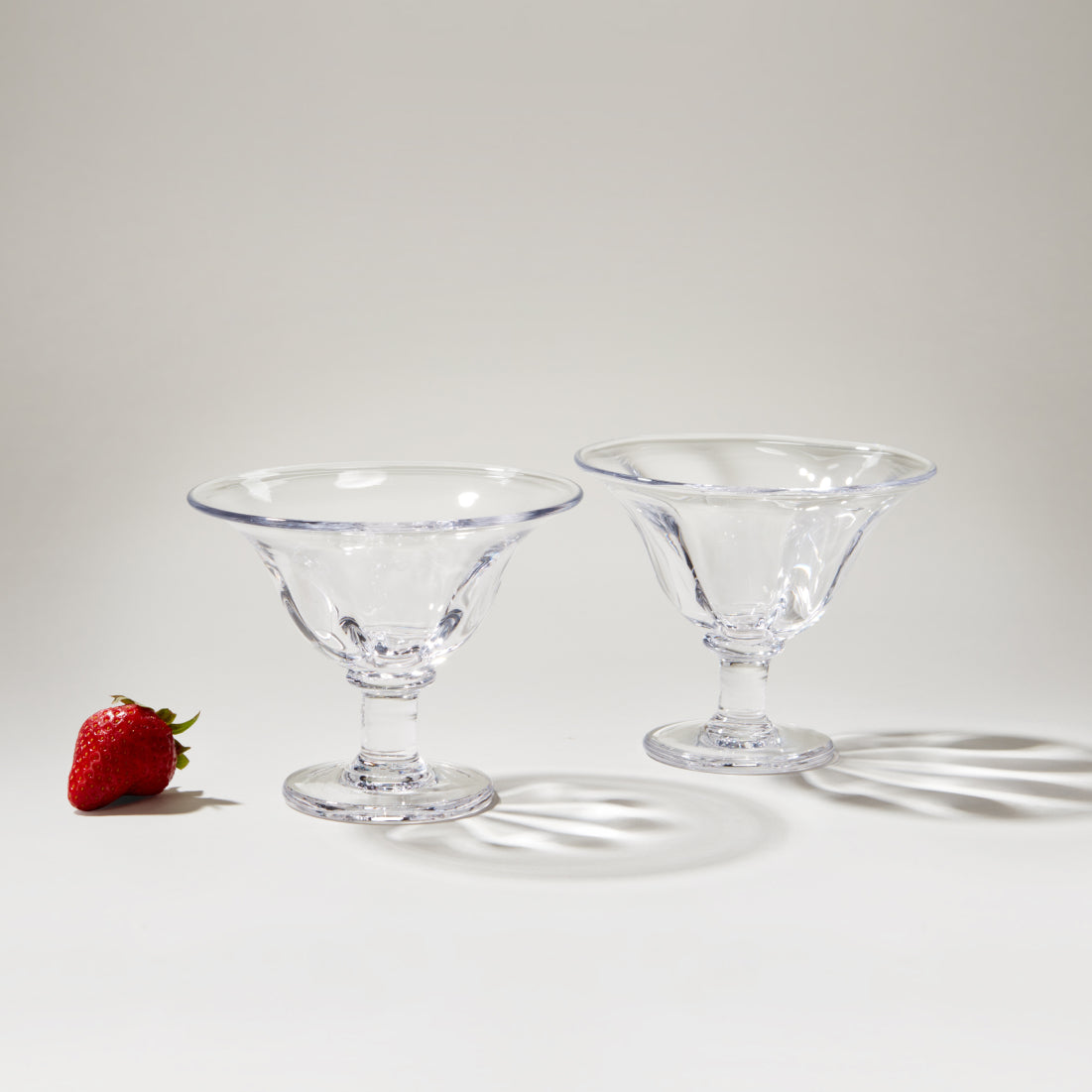 Two clear glass dessert bowls with pedestal bases and blue-tinted rims, displayed with a strawberry for scale.