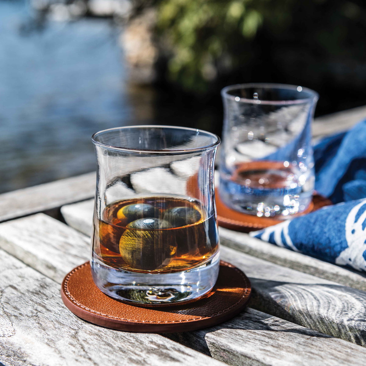 Two glasses of whiskey with spherical chilling stones on a wooden surface by water.