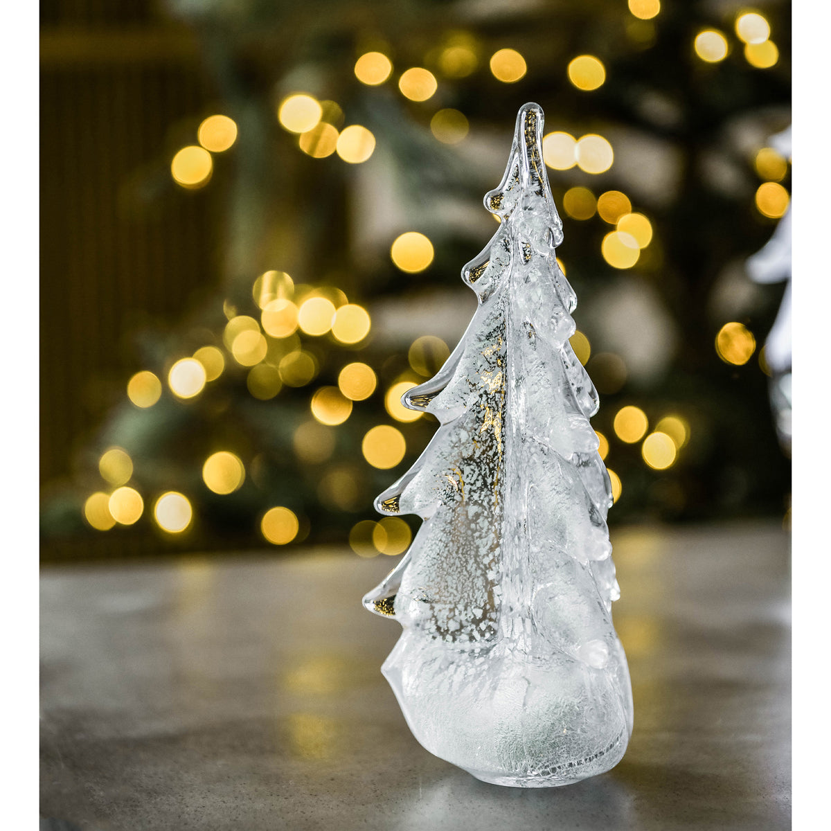 Frosted glass evergreen tree ornament with silver glitter detail and icicle coating against blurred golden lights.