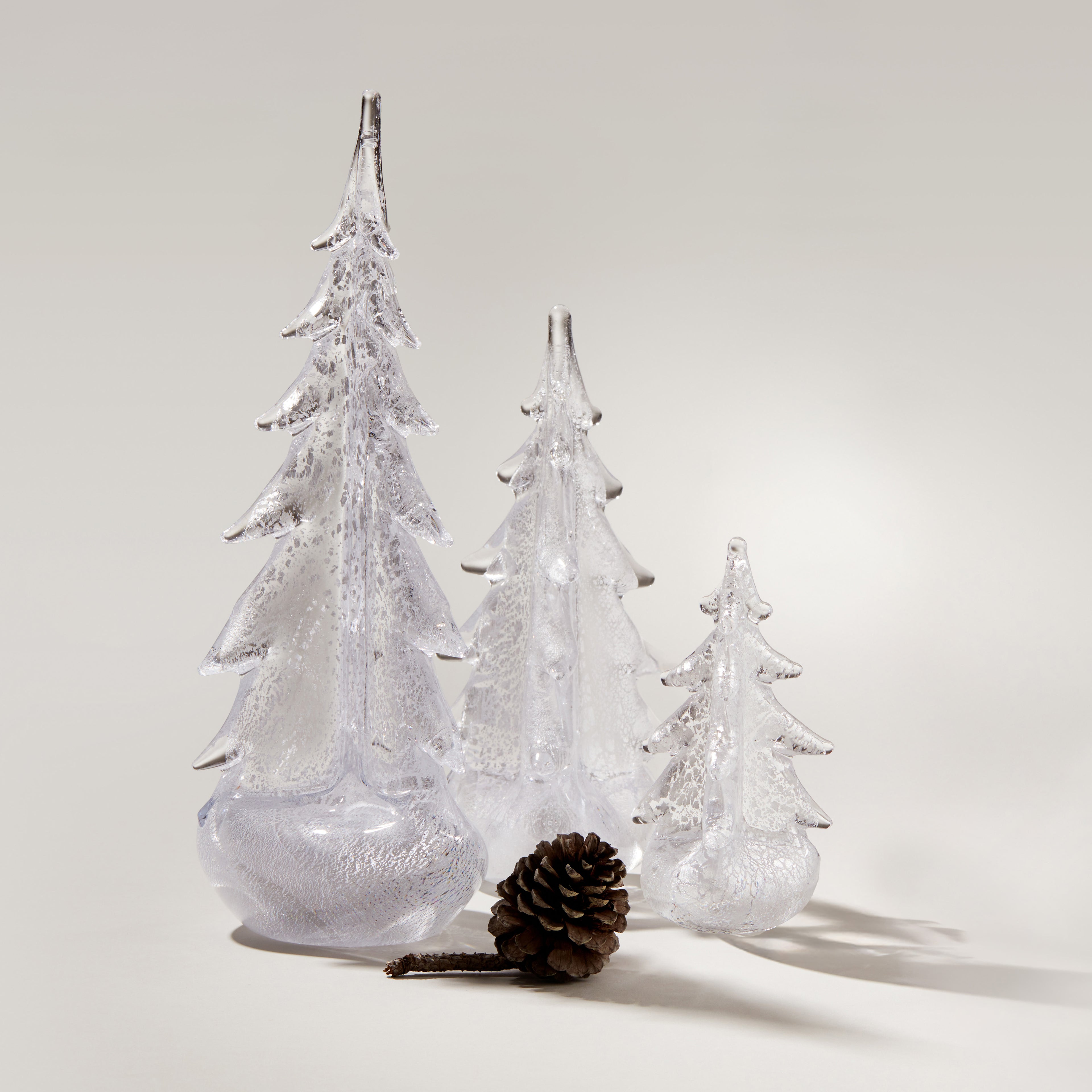 Three clear glass evergreen trees with frosted finish and a brown pinecone on neutral background.
