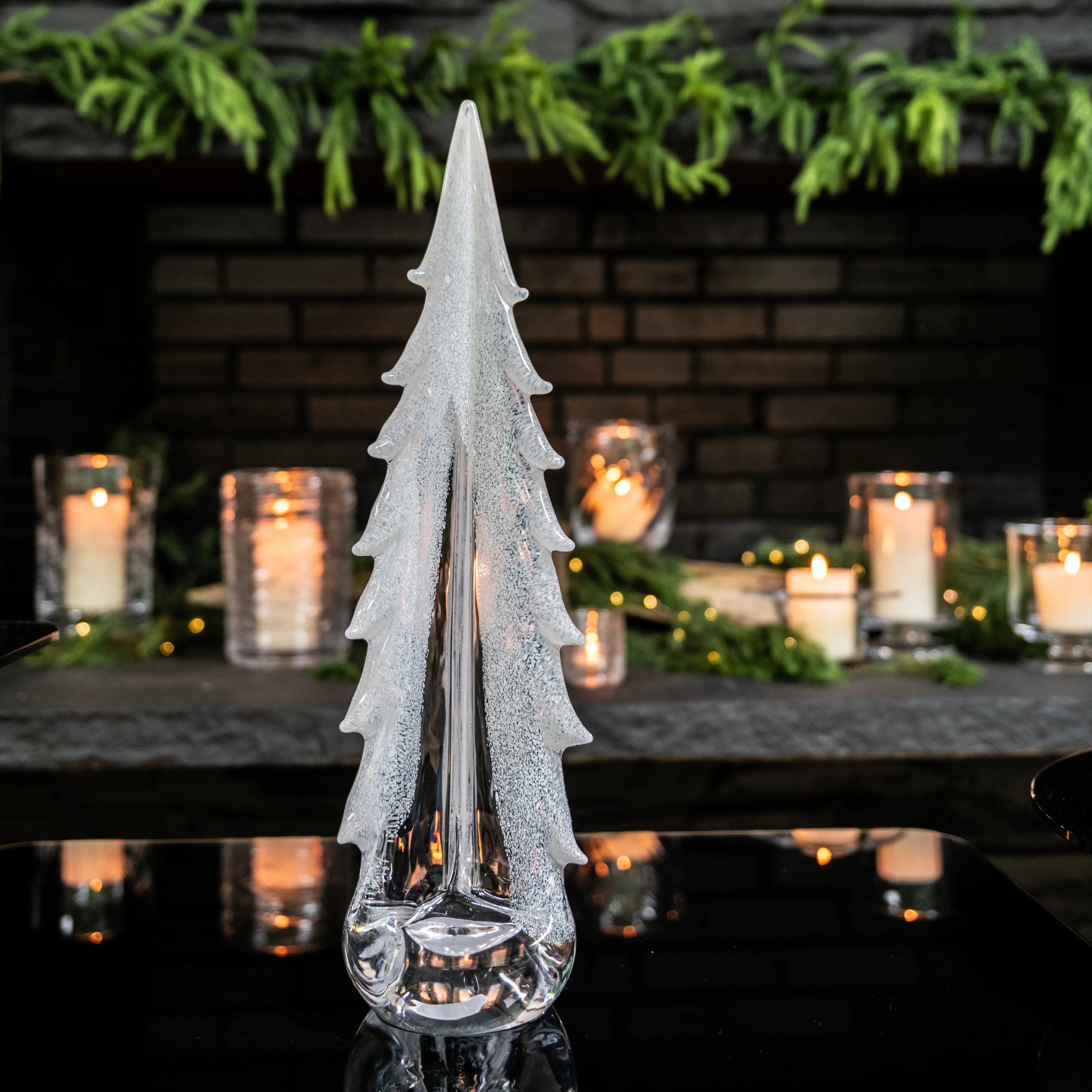 White frosted tree-shaped glass decor with textured finish and clear glass base, surrounded by candles and greenery.