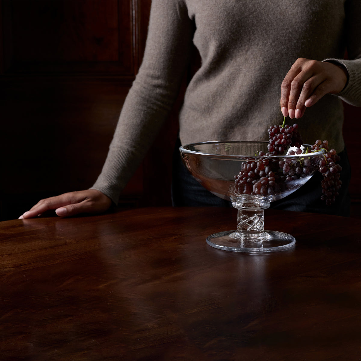 A clear glass pedestal bowl filled with dark red grapes sits on a polished dark wood table, with a person in a taupe sweater reaching in from the side.