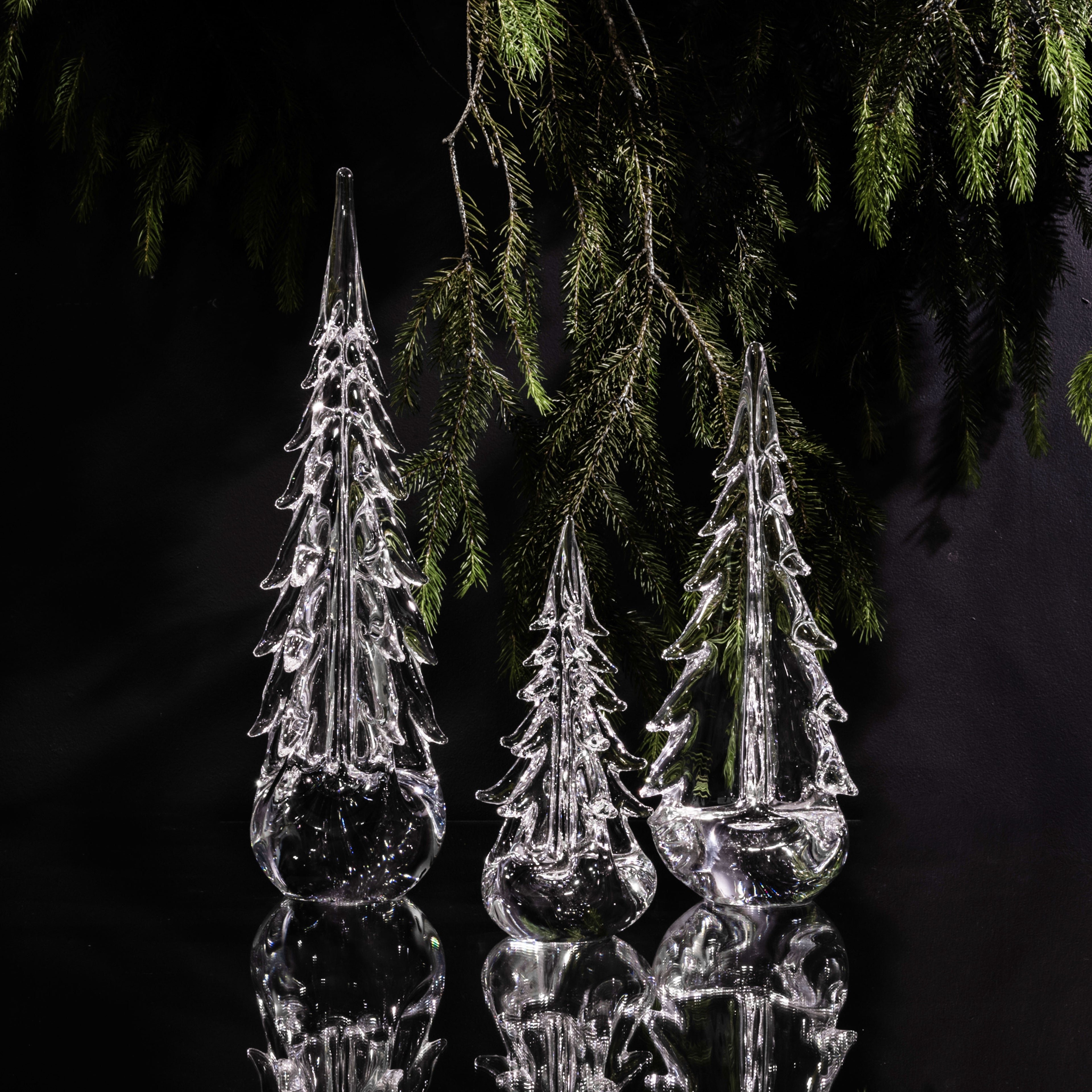 Three clear glass tree-shaped ornaments with evergreen branches suspended above against a black background.