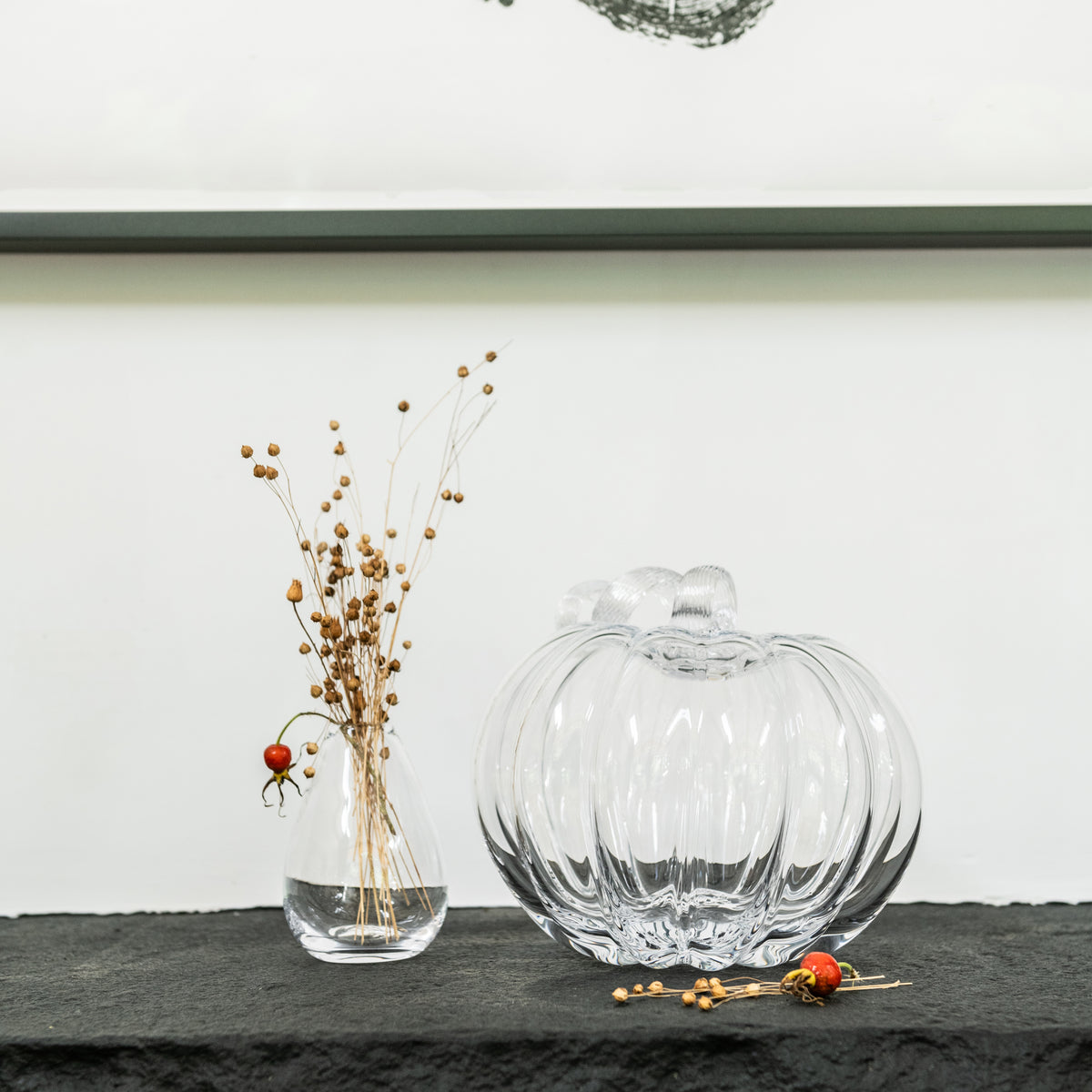Clear glass pumpkin-shaped decorative object with ribbed texture next to dried berry stems in a small vase.