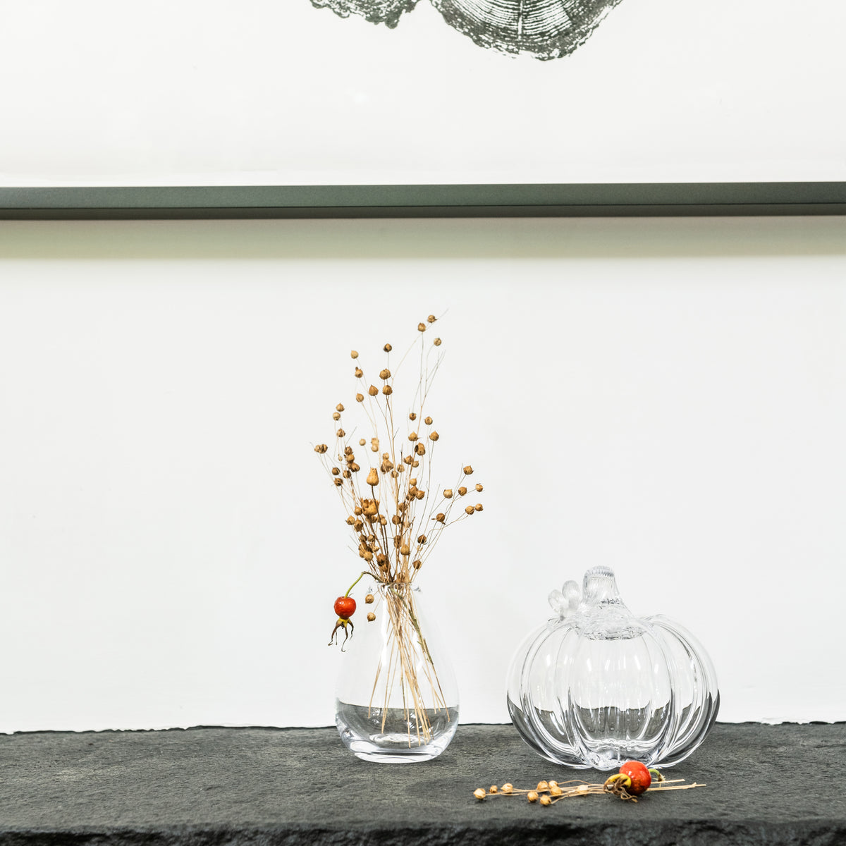Clear glass vase with dried golden berry stems and single red berry, alongside pumpkin-shaped glass ornament on dark shelf.