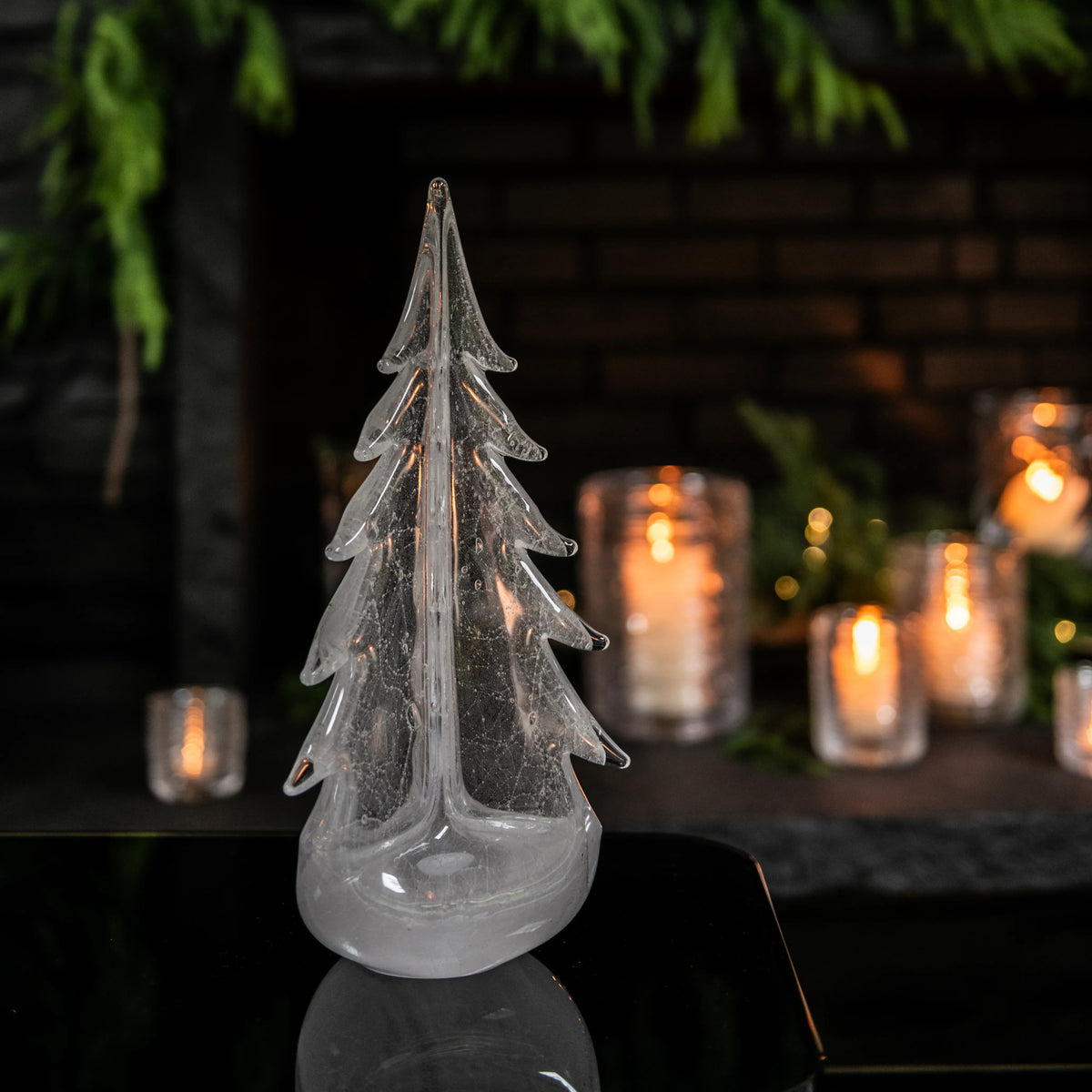 Clear glass evergreen tree candle with tiered branches and rounded base, displayed with lit candles and greenery in backgroun