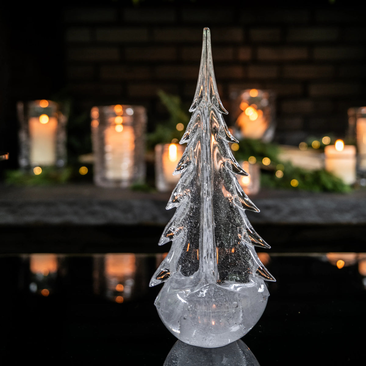 Clear glass tree-shaped candle with frosted finish and pointed top, displayed with lit candles blurred in background.