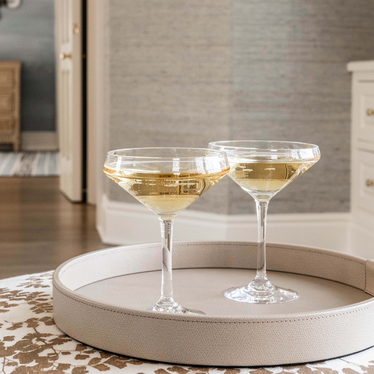 Two clear coupe glasses filled with pale wine on a cream-colored round tray.
