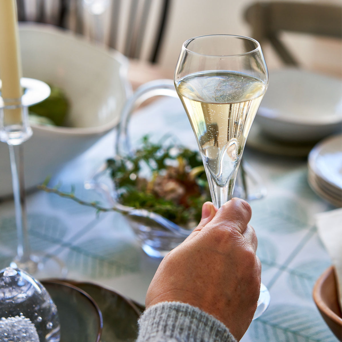 Hand holding a clear glass flute filled with sparkling wine at a dining table set with plates and tableware.