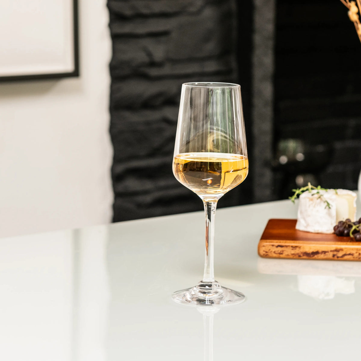 White wine in a stemmed glass on a white countertop with cheese and fruit on a wooden board in the background.