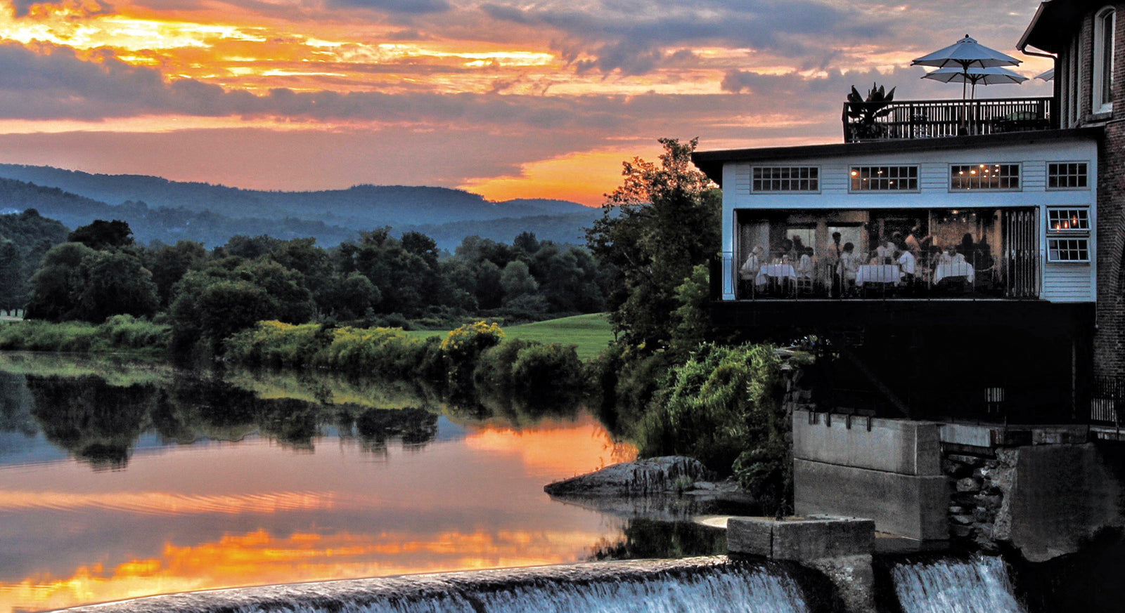 The Simon Pearce Mill in Quechee, Vermont at dusk, illuminated from within as guests dine on the terrace above the Ottauquechee River waterfall, with the historic brick mill building at sunset