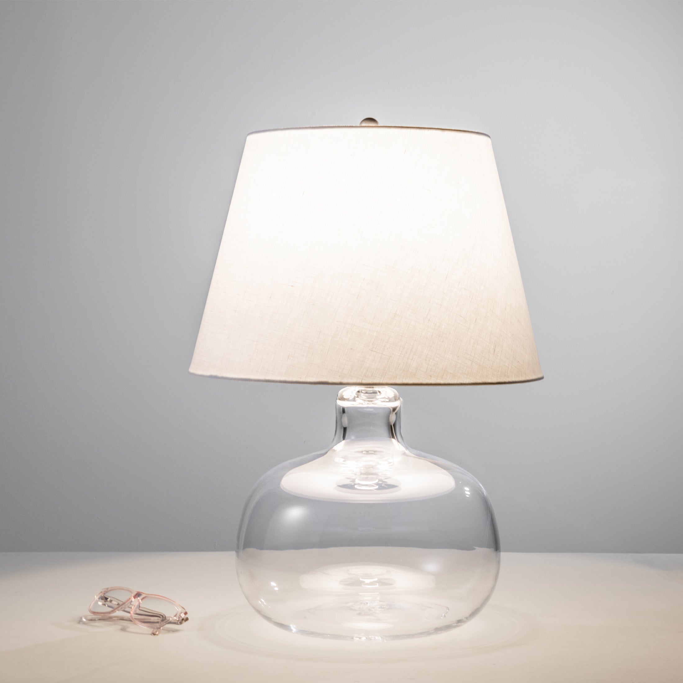 Clear glass round base lamp with cream linen tapered shade and metal finial.