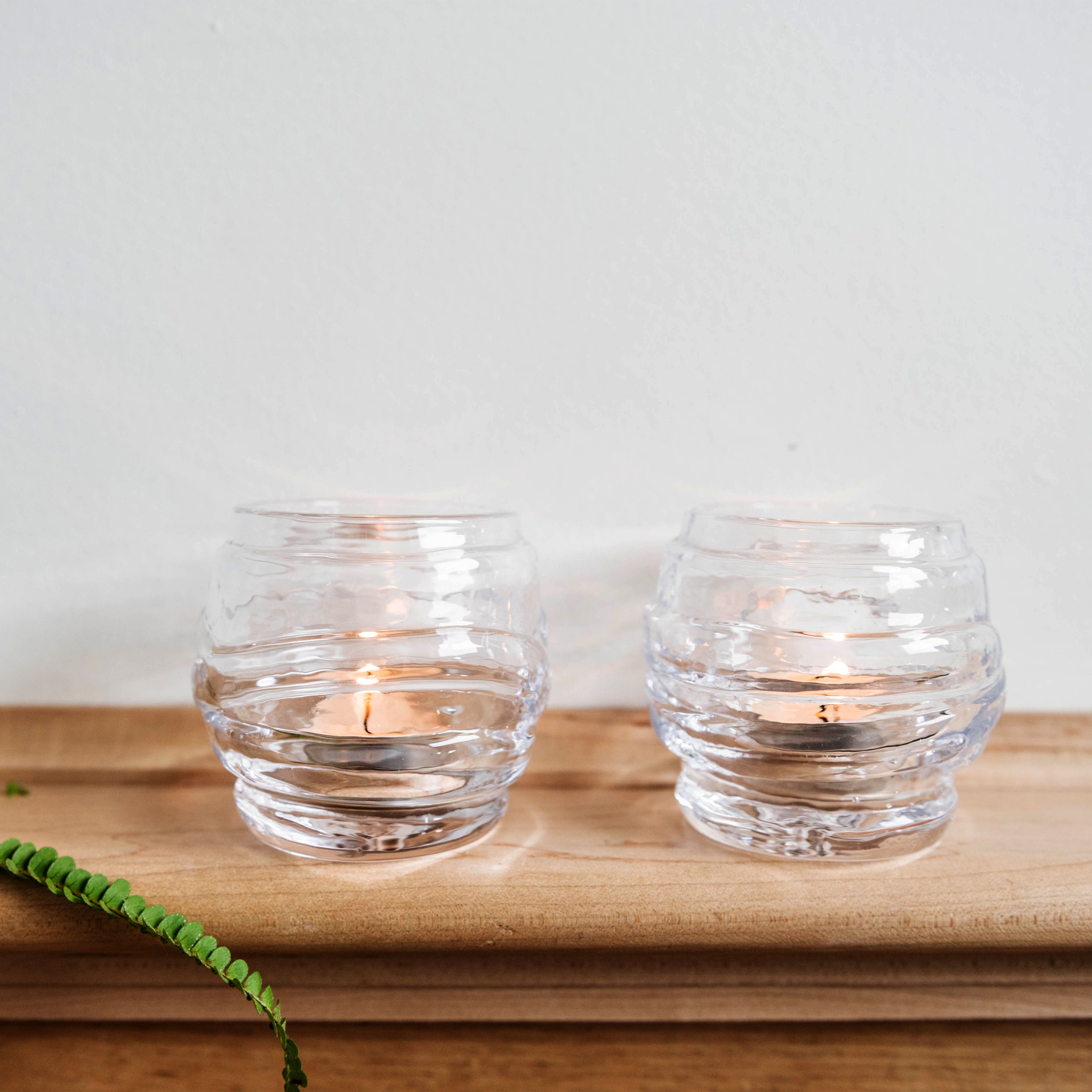 Two clear glass tealight holders with horizontal ribbed pattern, one containing a lit candle, displayed on wooden tray.