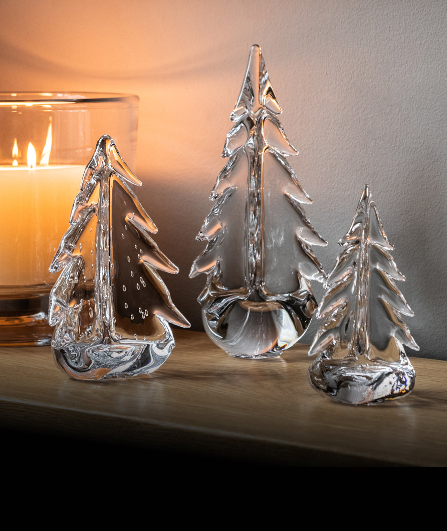 Three clear glass Christmas trees on a wooden surface beside a lit candle