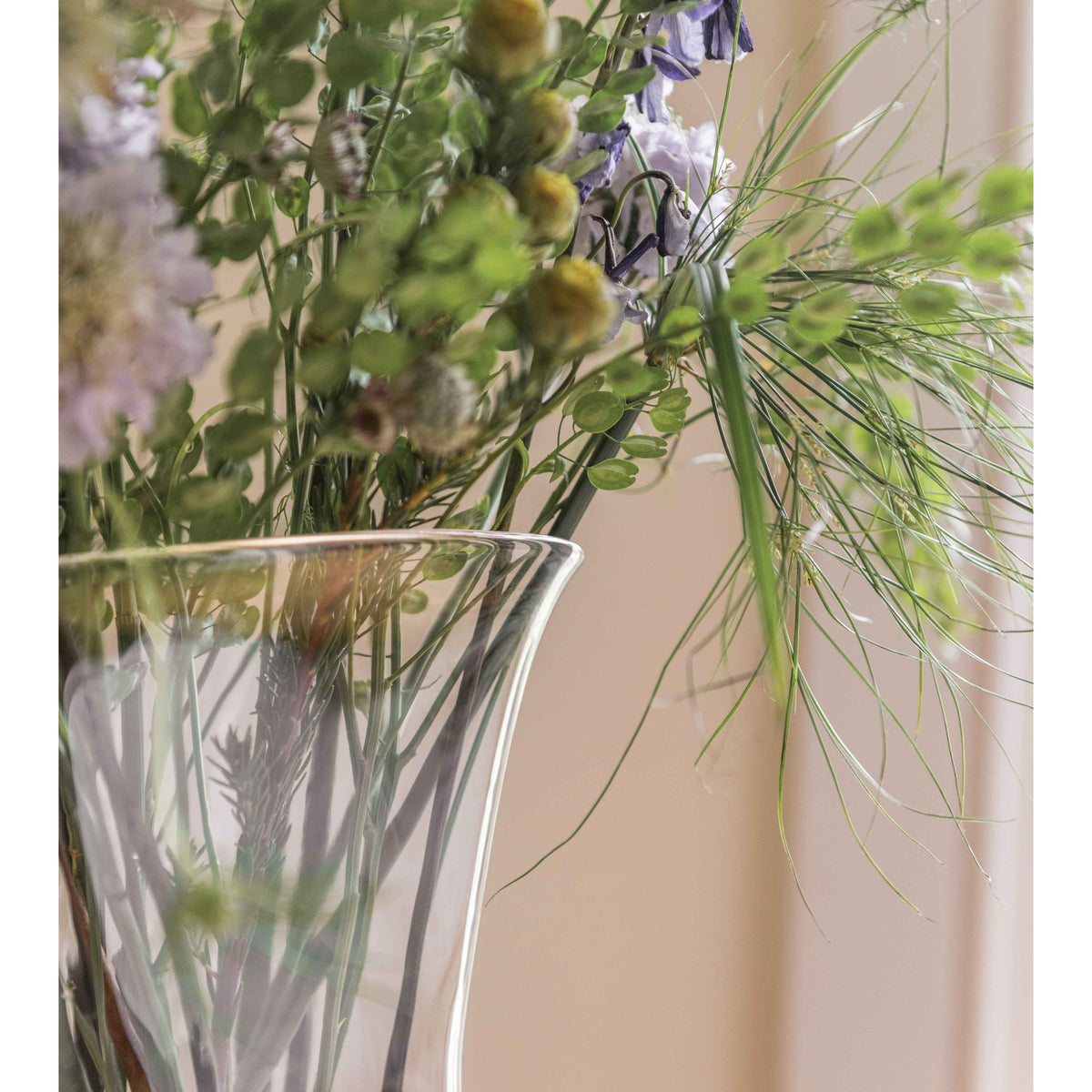 Mixed flower arrangement in clear glass vase with purple blooms, green seed pods, and delicate filler stems against neutral b