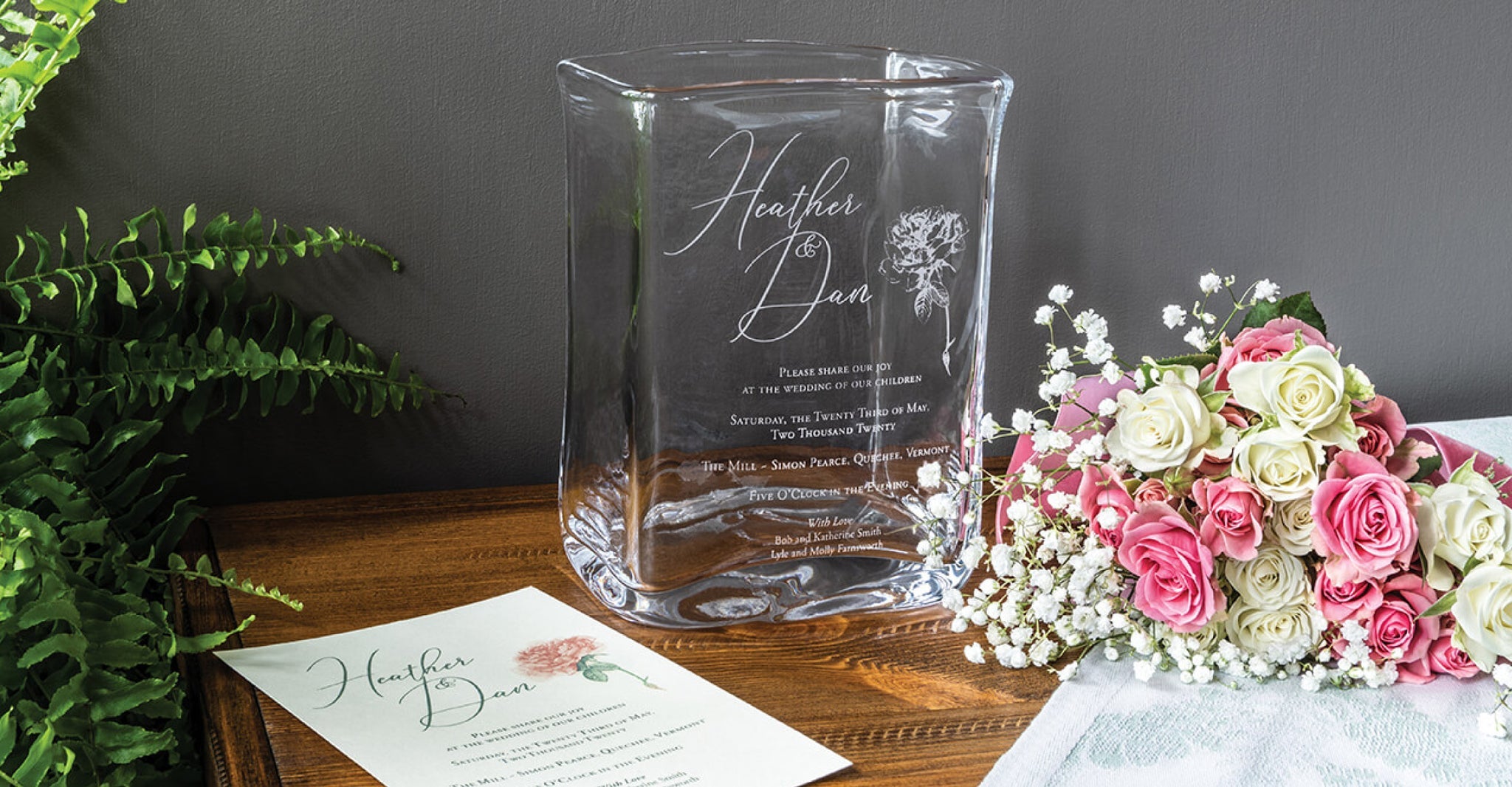 Clear glass vase with engraved wedding names and details, displayed with pink and white roses and wedding invitation.