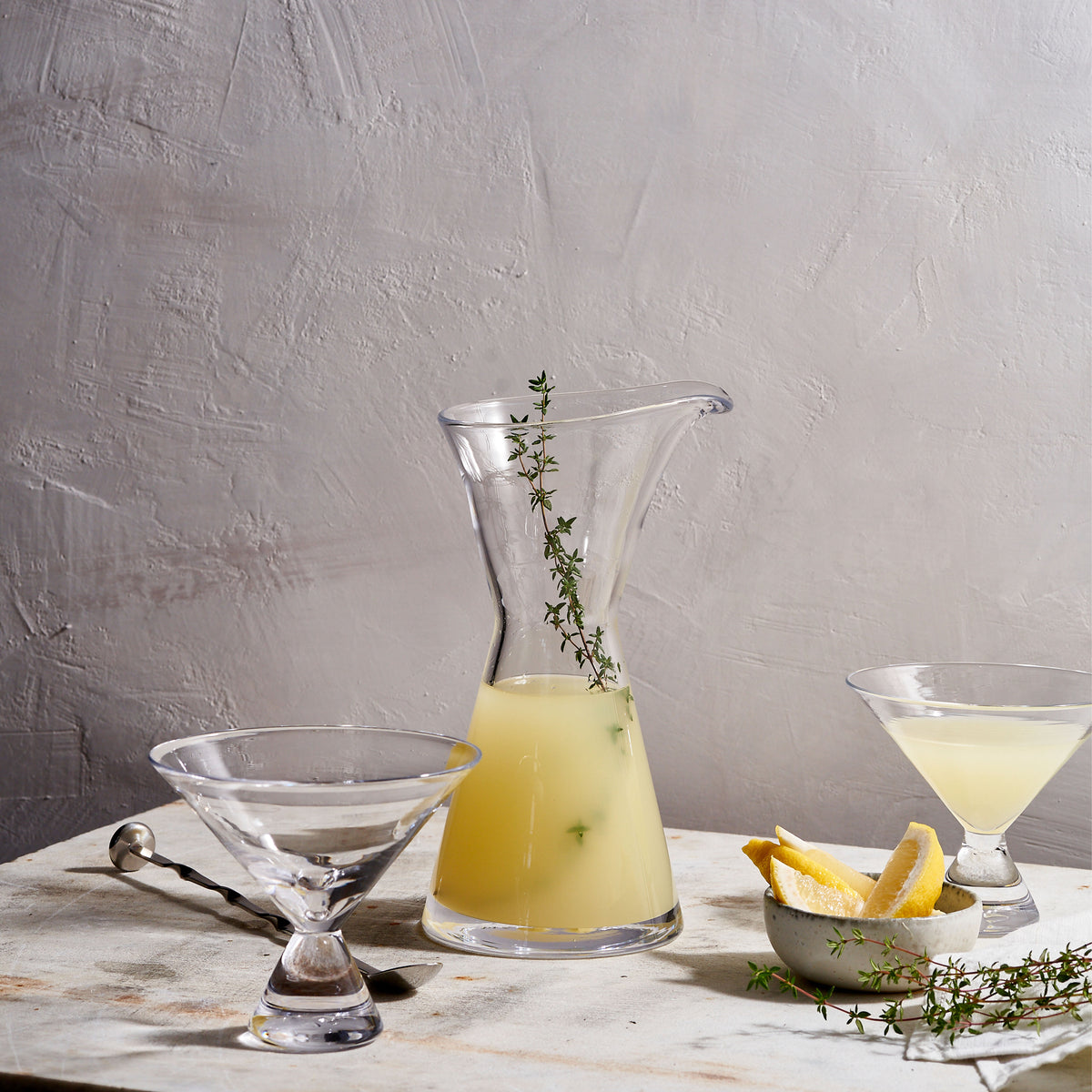 Glass pitcher with yellow cocktail and fresh thyme, two martini glasses, bar spoon, lemon peels, and herbs on marble surface.