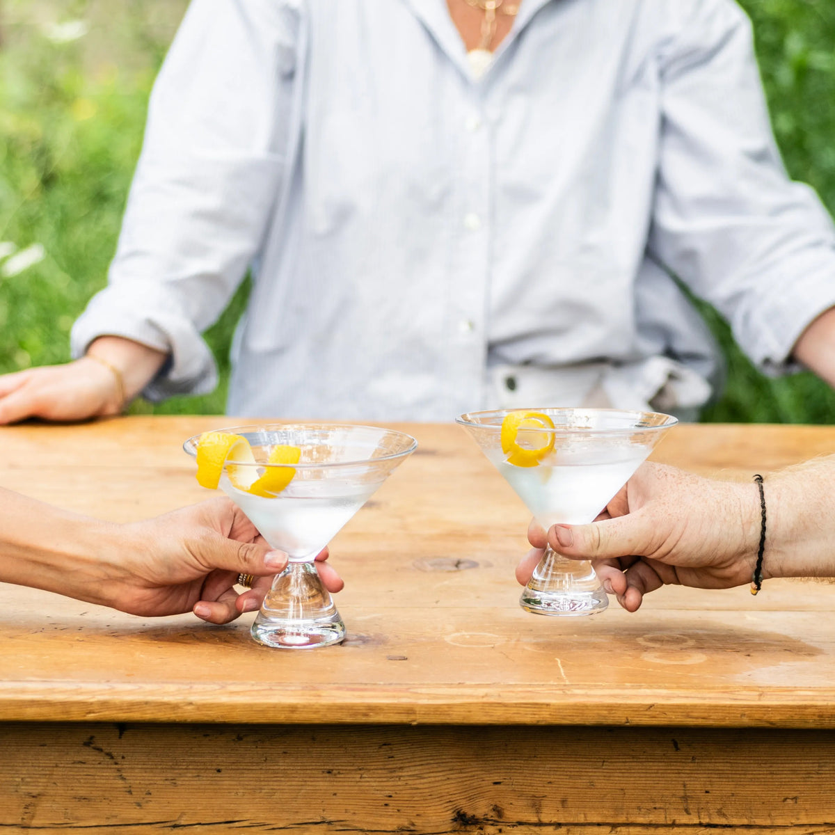 Two clear stemless martini glasses filled with white liquid and lemon twists, held by two people at a wooden table.