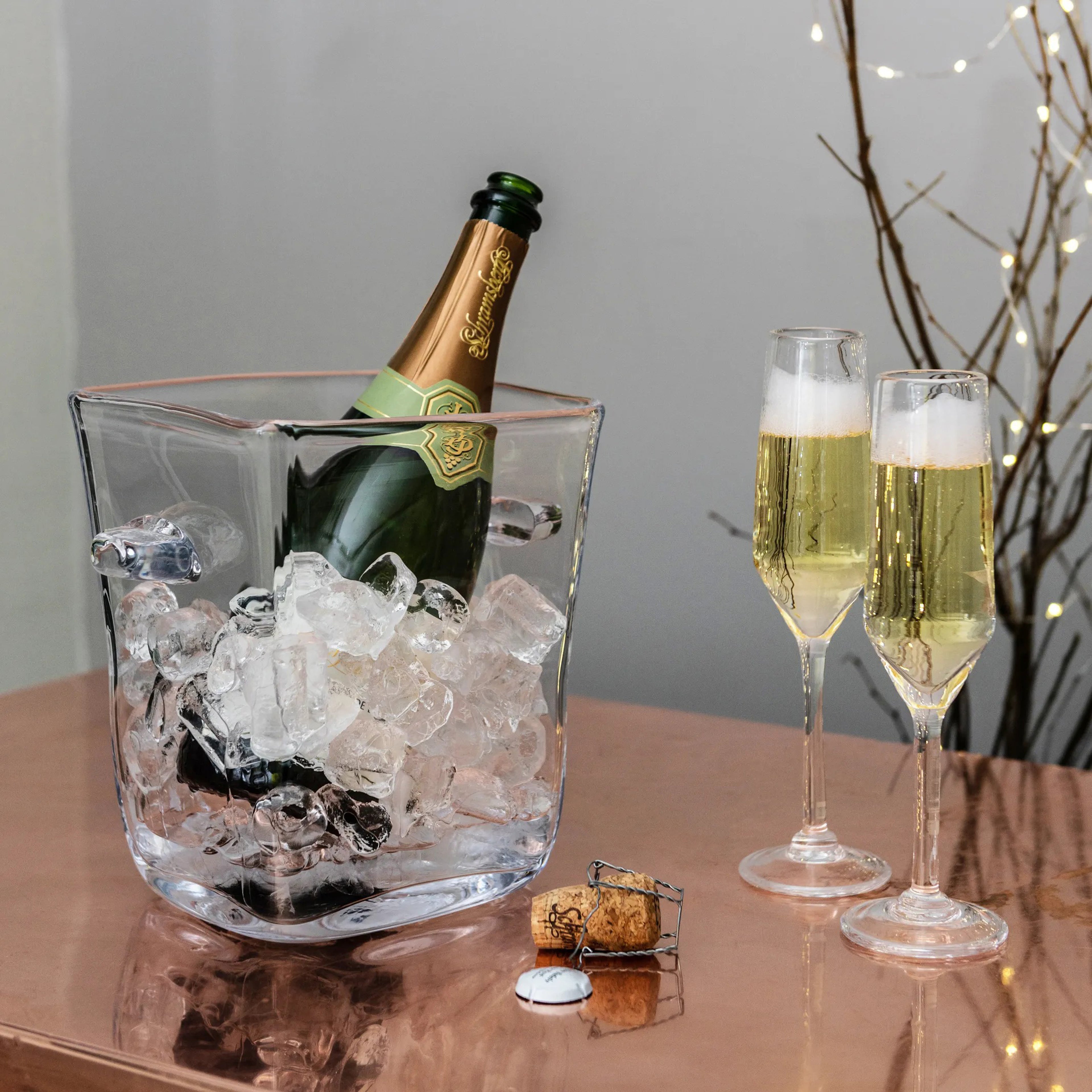 Clear glass ice bucket holding a champagne bottle surrounded by ice, with two filled champagne flutes beside it.