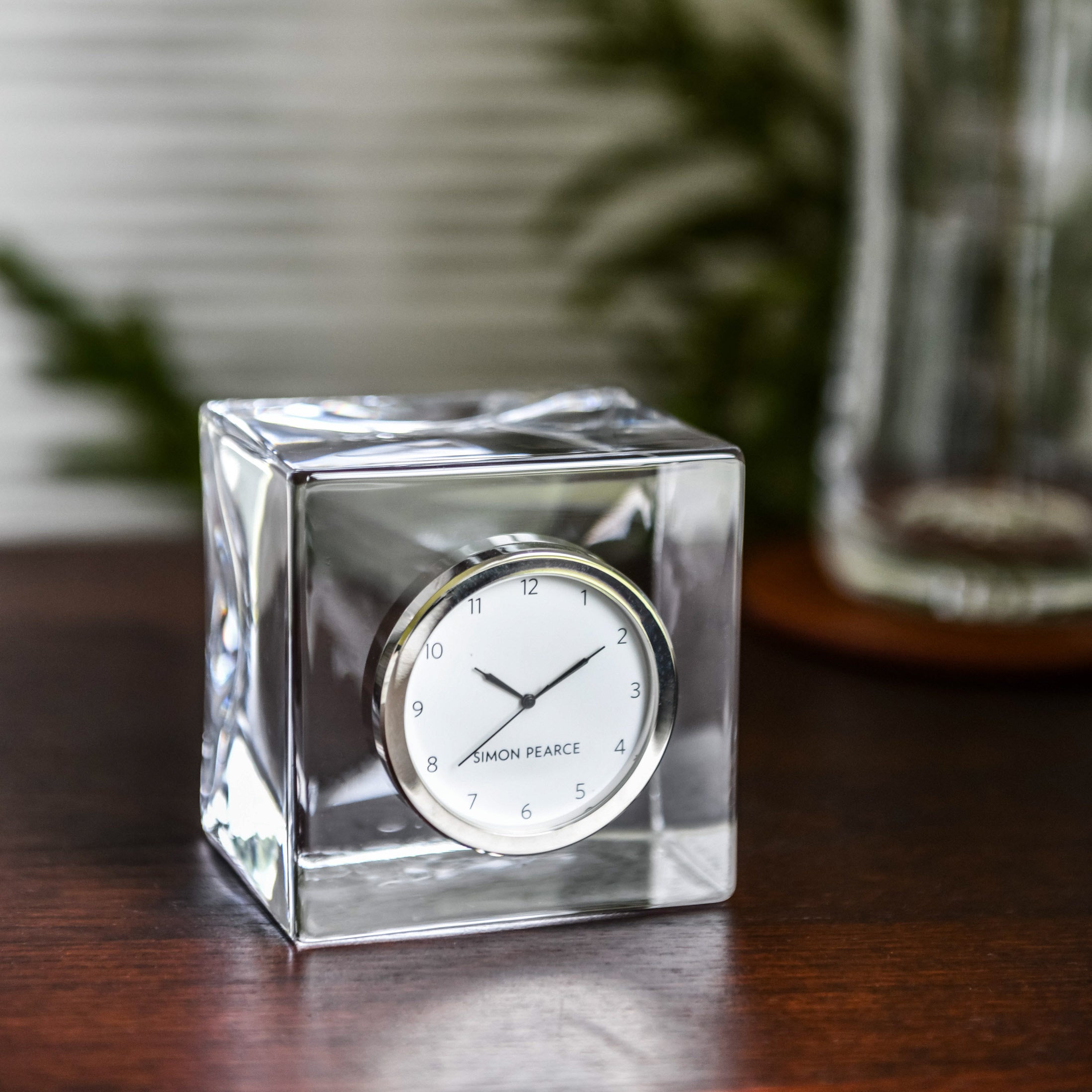 Clear glass cube desk clock with white dial, black hands, and silver trim on wooden surface.