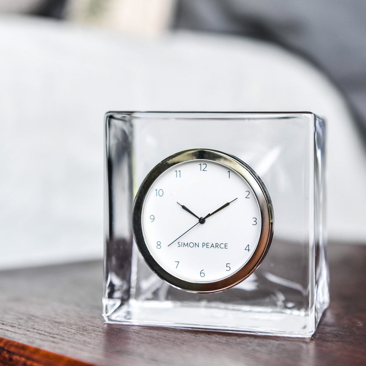 Round clock with white face and black hands mounted in clear glass square case on wooden surface.
