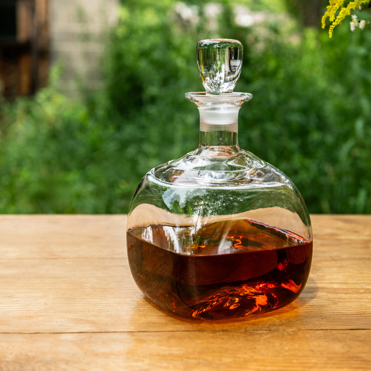 Clear glass decanter with rounded base, narrow neck, and stopper filled with amber liquid on wooden surface.