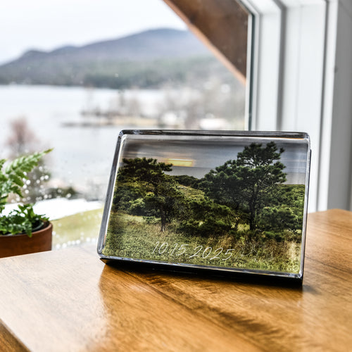 Horizontal acrylic photo block displaying landscape image of trees and water, displayed on wooden table near window.
