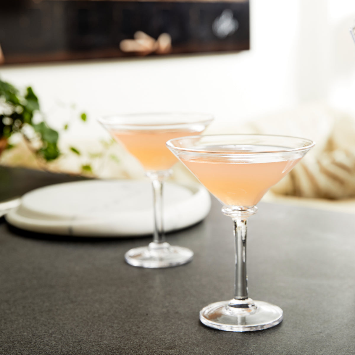 Two martini glasses filled with pale pink cocktail, displayed on a gray countertop with place settings.