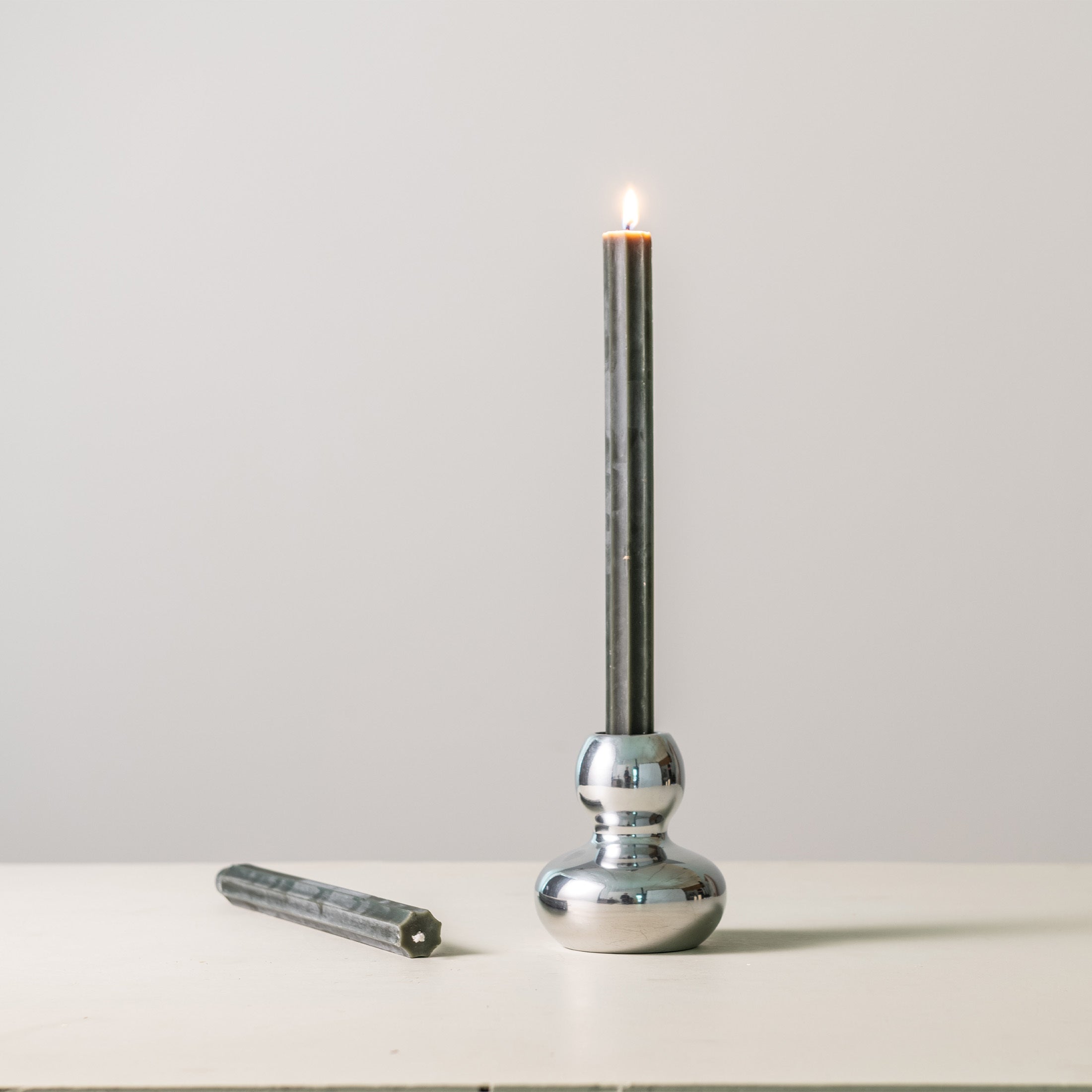 Polished silver aluminum candlestick with rounded base holding a lit taper candle, shown with extinguisher tool.