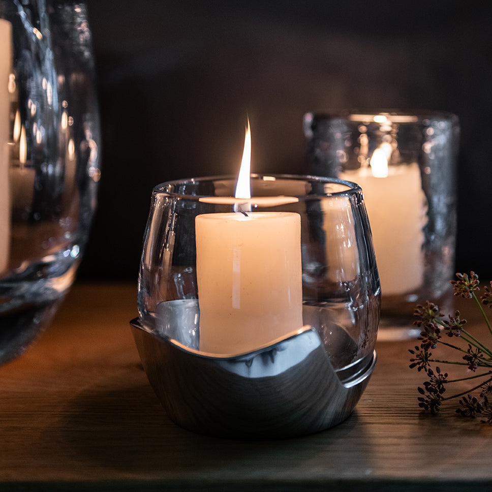Lit candle in clear glass holder with black brushstroke design, surrounded by dried botanicals and additional candles on wood