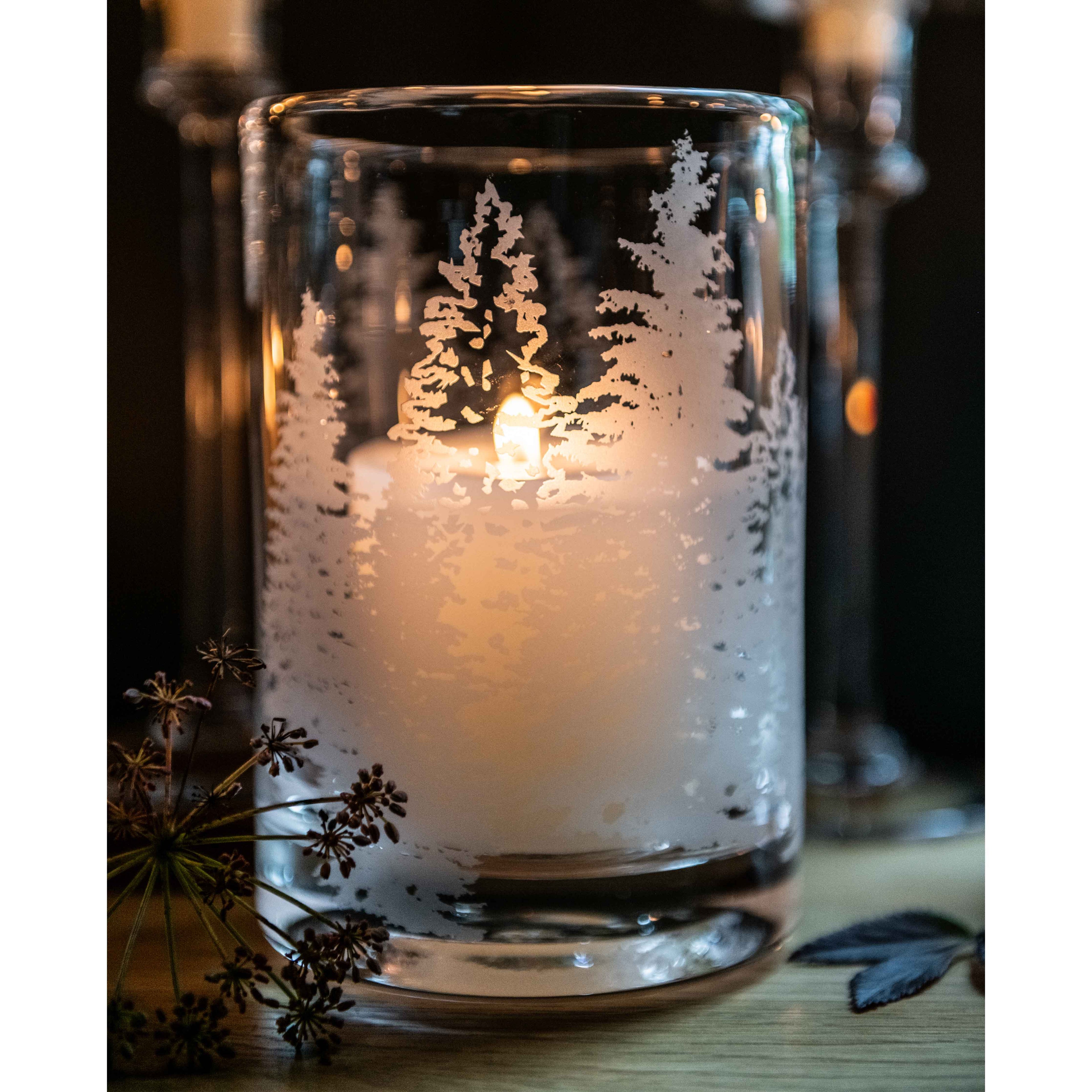 Clear glass hurricane with white engraved evergreen trees and lit candle inside.